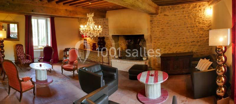 Salon chaleureux avec murs en pierre, cheminée, lustre, fauteuils variés et tables basses dans un cadre rustique.