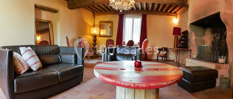 Salon rustique avec poutres apparentes, cheminée, mobilier en cuir et table basse en bois rouge dans un cadre chaleureux.