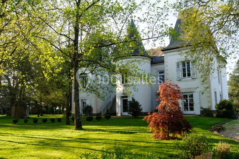 Grande maison blanche avec tourelles, entourée d'arbres et d'un jardin verdoyant sous une lumière ensoleillée.