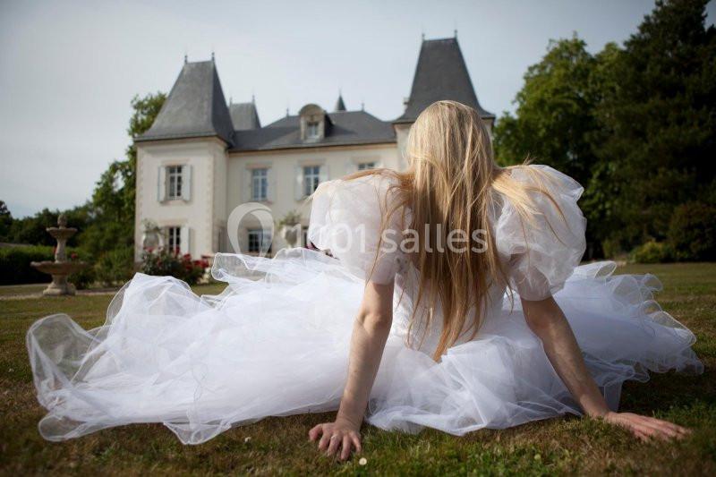 Femme en robe blanche volumineuse assise sur l'herbe, face à un manoir entouré de verdure.