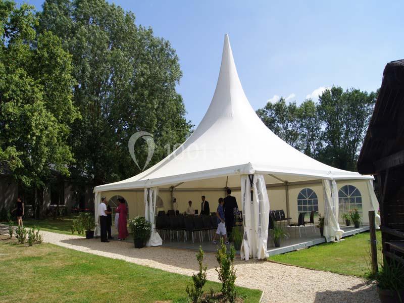 Tente blanche de réception montée dans un jardin, entourée d'arbres, avec des personnes à l'entrée.
