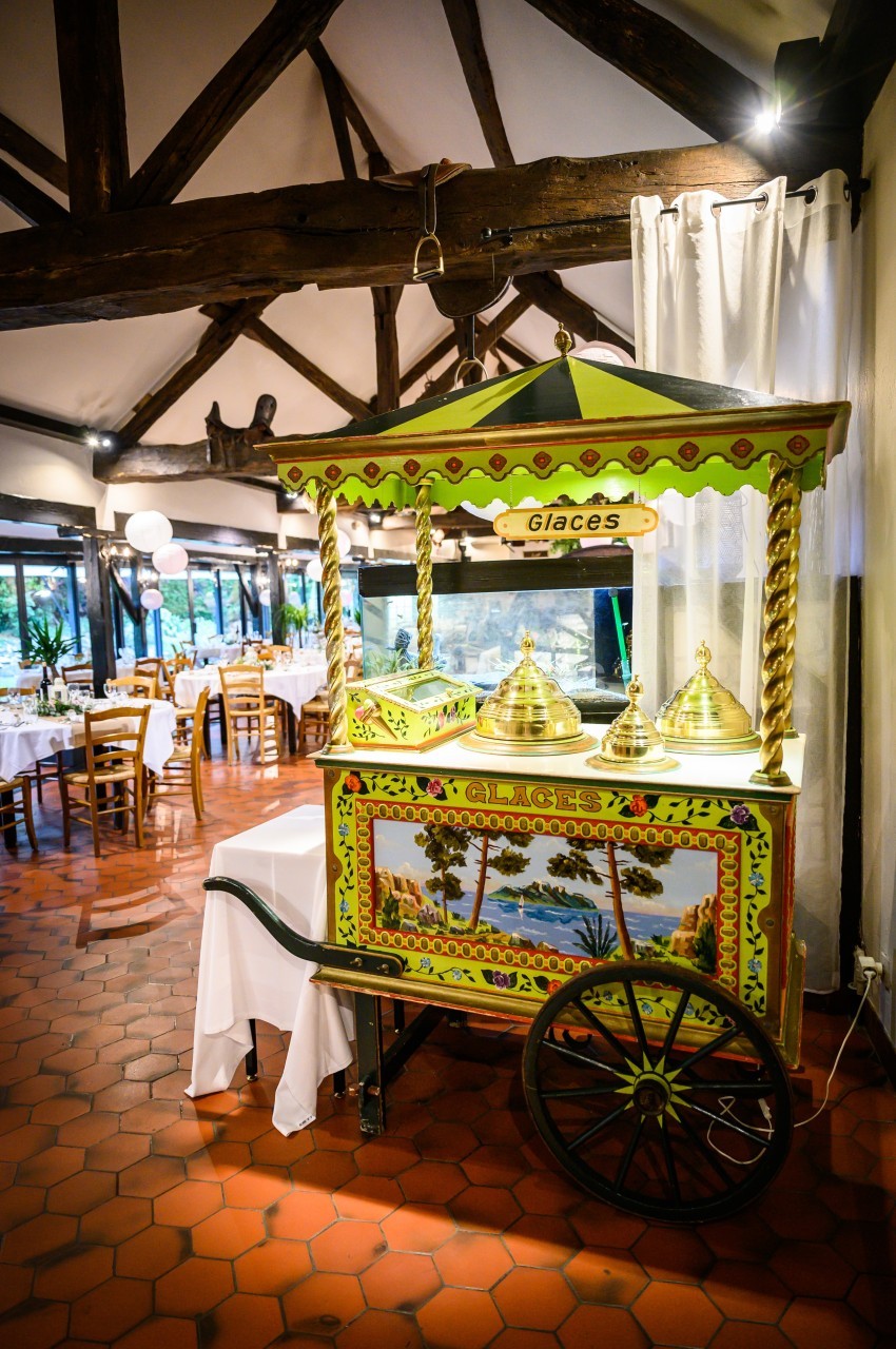 Chariot de glaces coloré avec couvercles dorés, installé dans une salle de réception au sol en carreaux rouges.