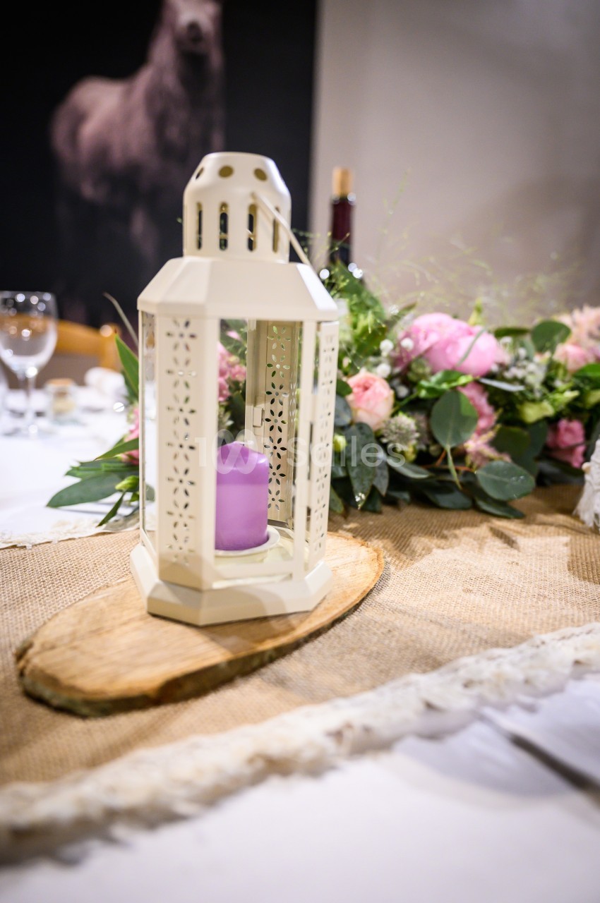 Lanterne blanche avec une bougie violette sur une table décorée de fleurs et d'un chemin de table en jute.