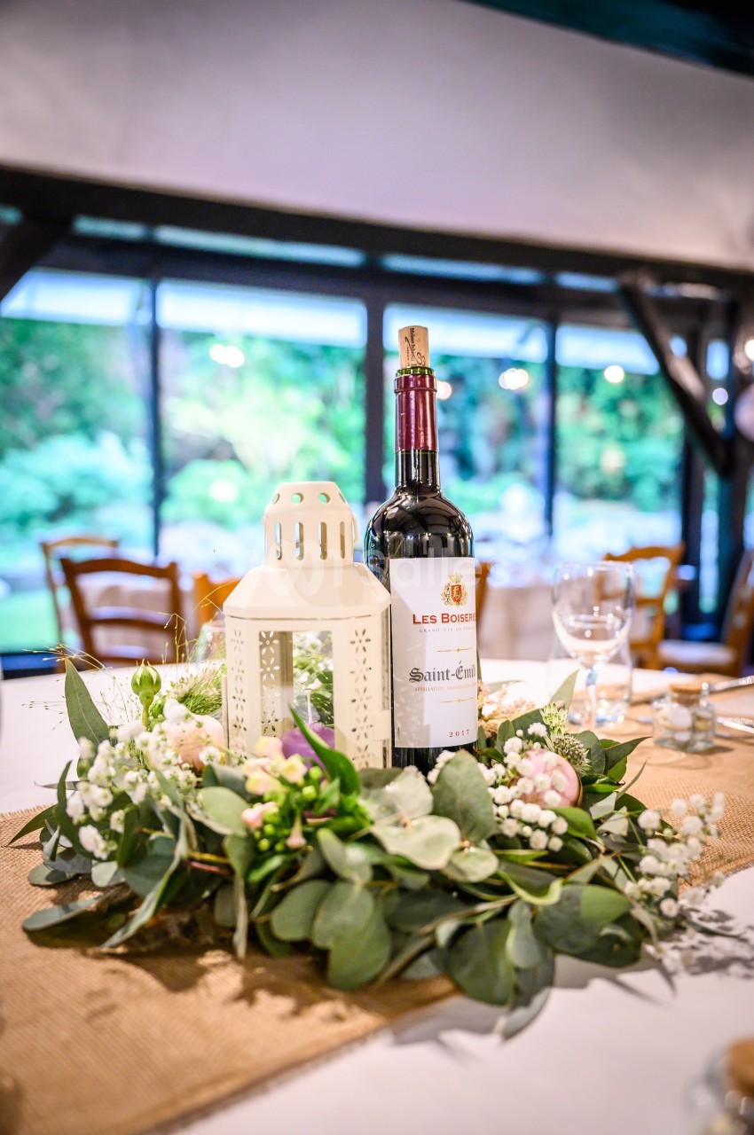 Bouteille de vin rouge posée sur une table décorée de feuillage, fleurs blanches et une lanterne blanche.