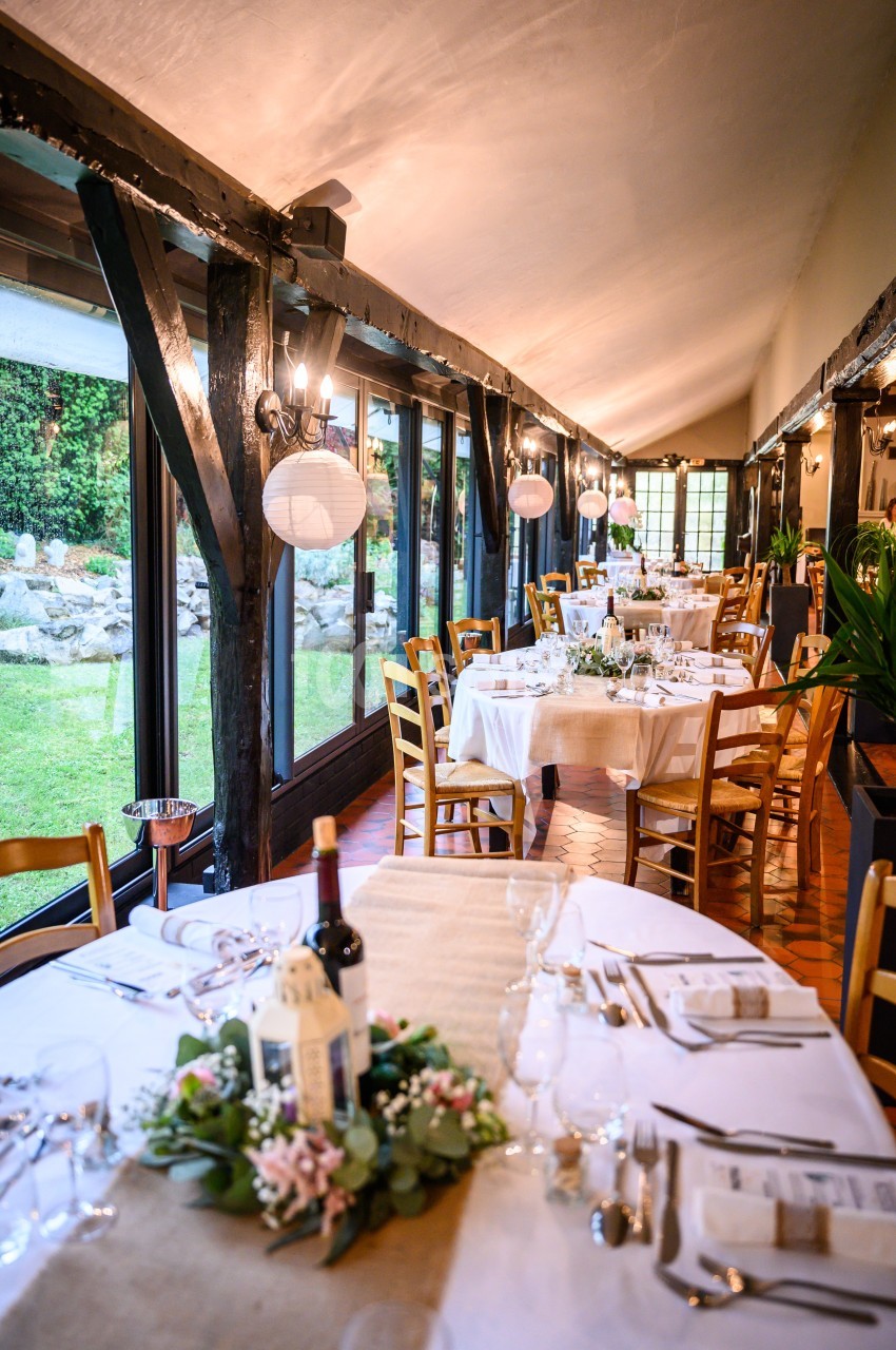 Salle de réception lumineuse avec tables décorées, nappes blanches, chaises en bois et vue sur un jardin extérieur.