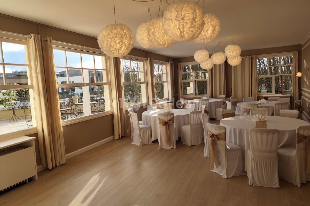 Salle lumineuse avec grandes fenêtres, tables rondes dressées de nappes blanches et chaises décorées de nœuds dorés.