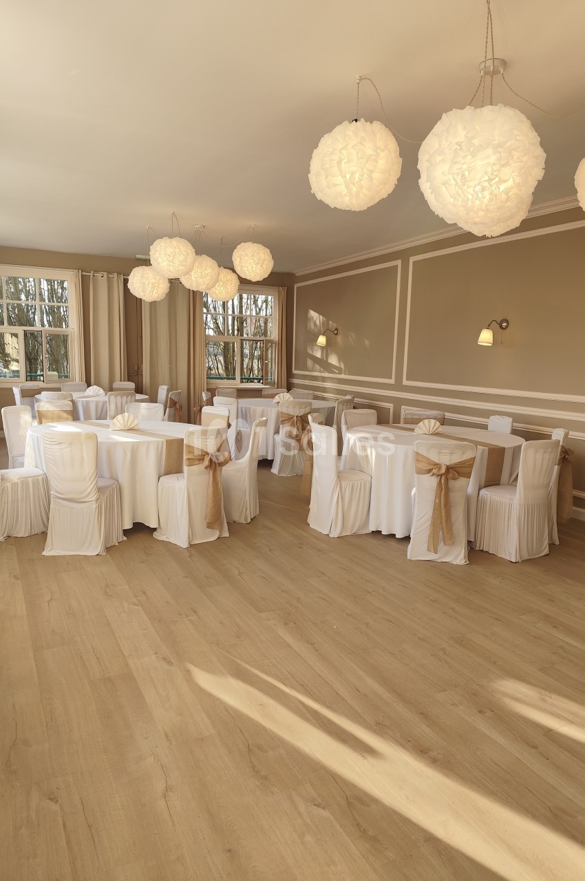 Salle de réception lumineuse avec tables rondes dressées, chaises blanches ornées de nœuds dorés et suspensions décoratives.