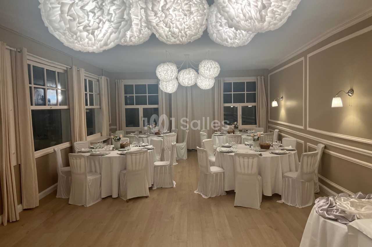 Salle de réception élégante avec tables rondes dressées, chaises blanches et suspensions lumineuses décoratives.