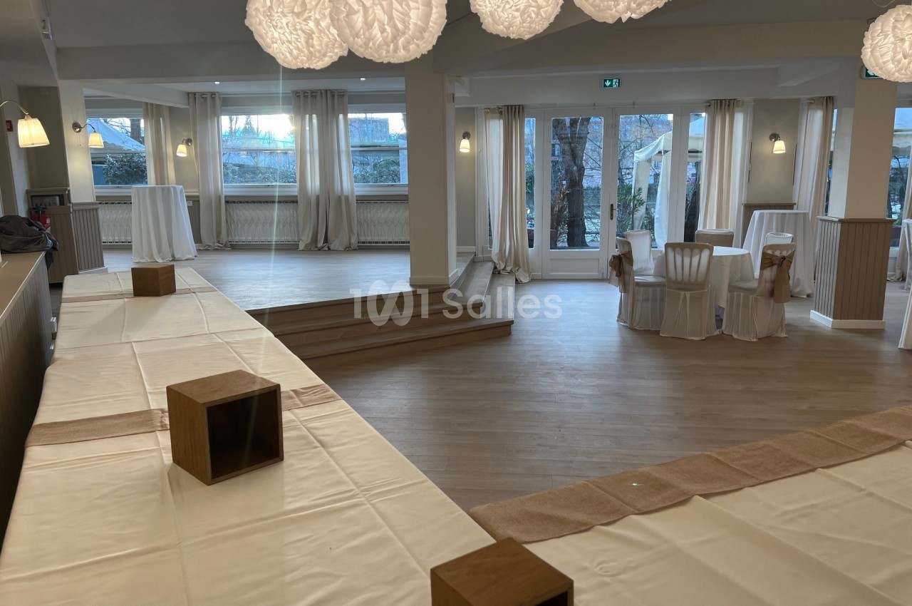 Salle lumineuse avec parquet, tables et chaises recouvertes de housses blanches, décorée de suspensions en papier.