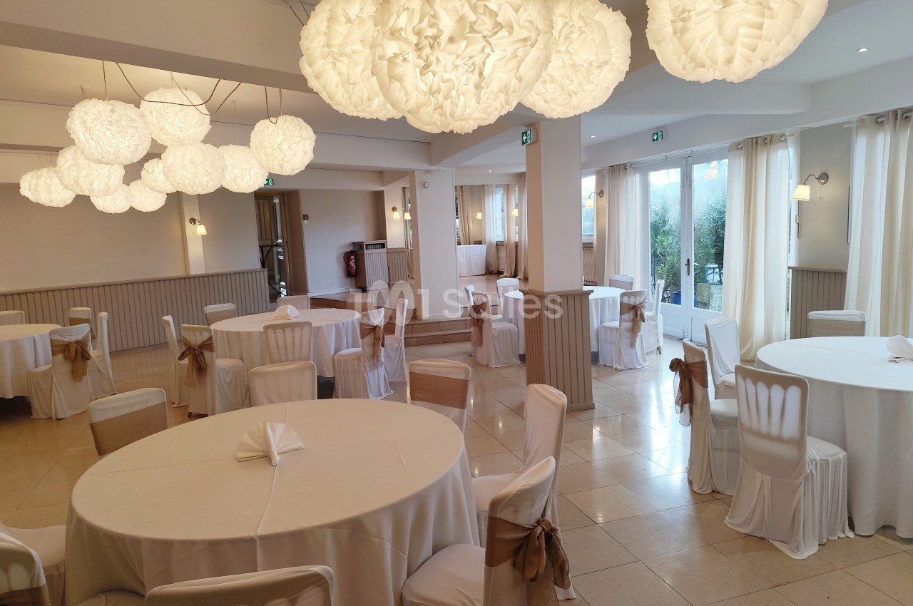 Salle de réception lumineuse avec tables rondes dressées de nappes blanches et décorations suspendues en papier blanc.