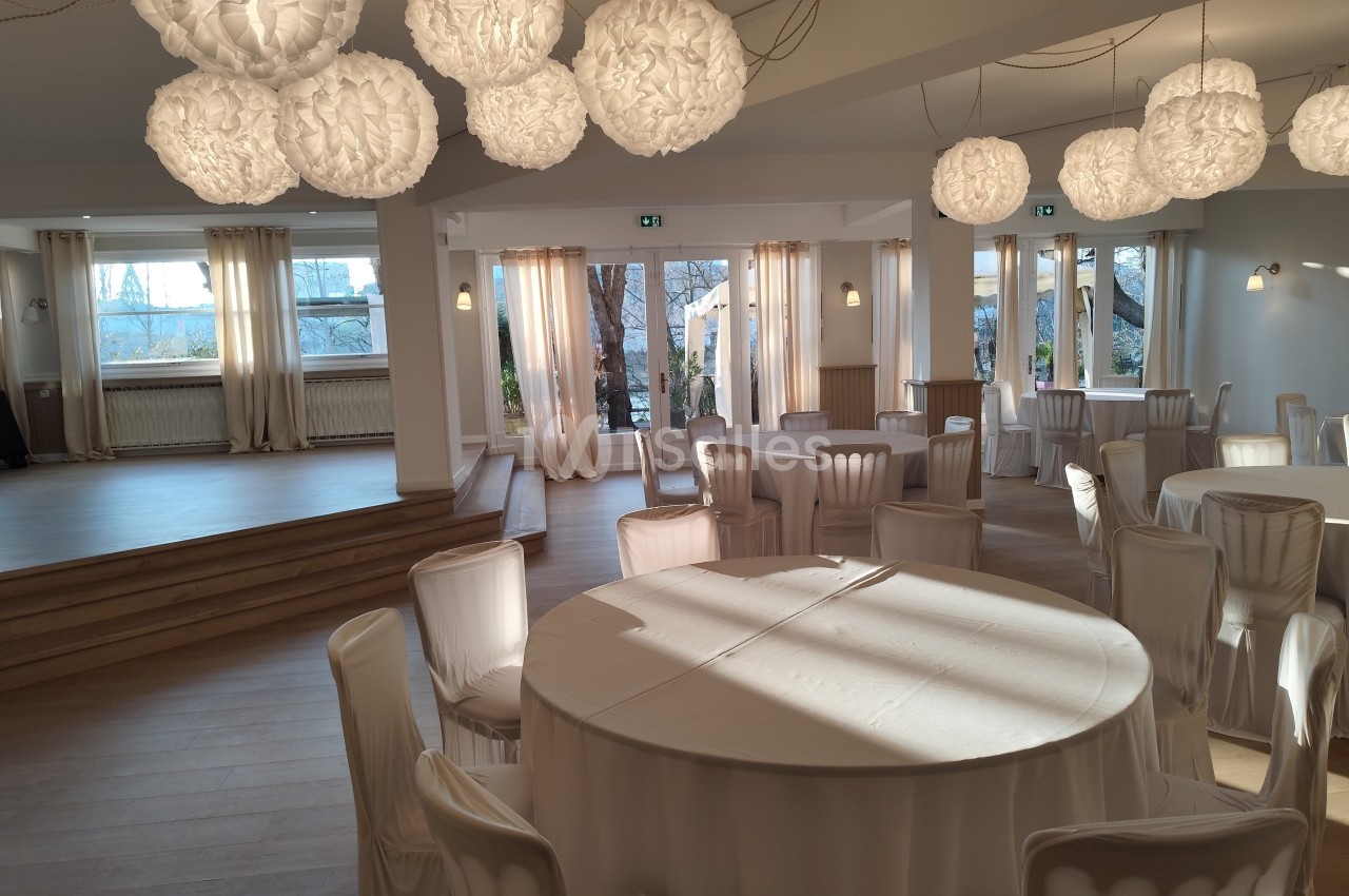Salle de réception lumineuse avec tables rondes nappées de blanc, éclairée par des suspensions décoratives.