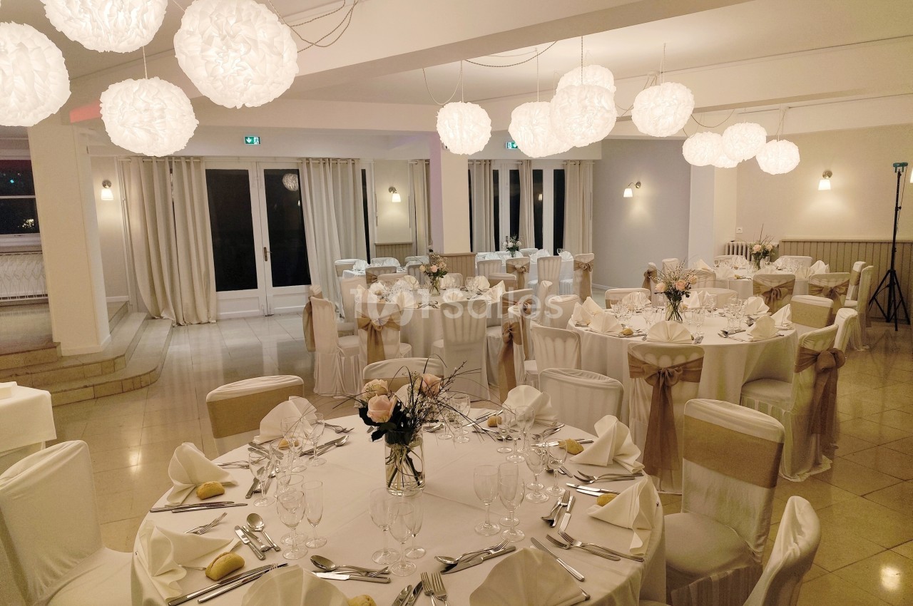 Salle de réception élégamment décorée avec des tables rondes dressées, des chaises habillées et des luminaires suspendus.
