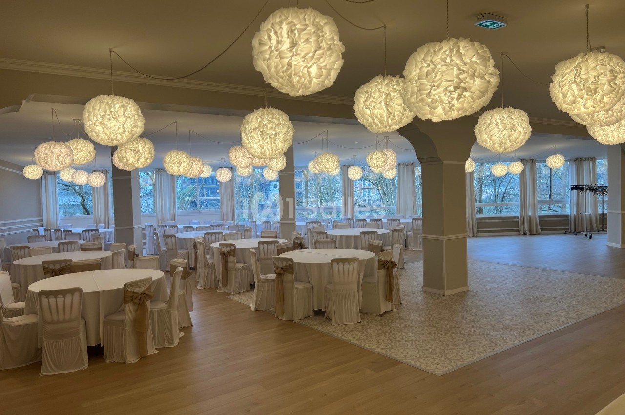 Salle de réception lumineuse avec tables rondes dressées de nappes blanches et suspensions décoratives en papier.