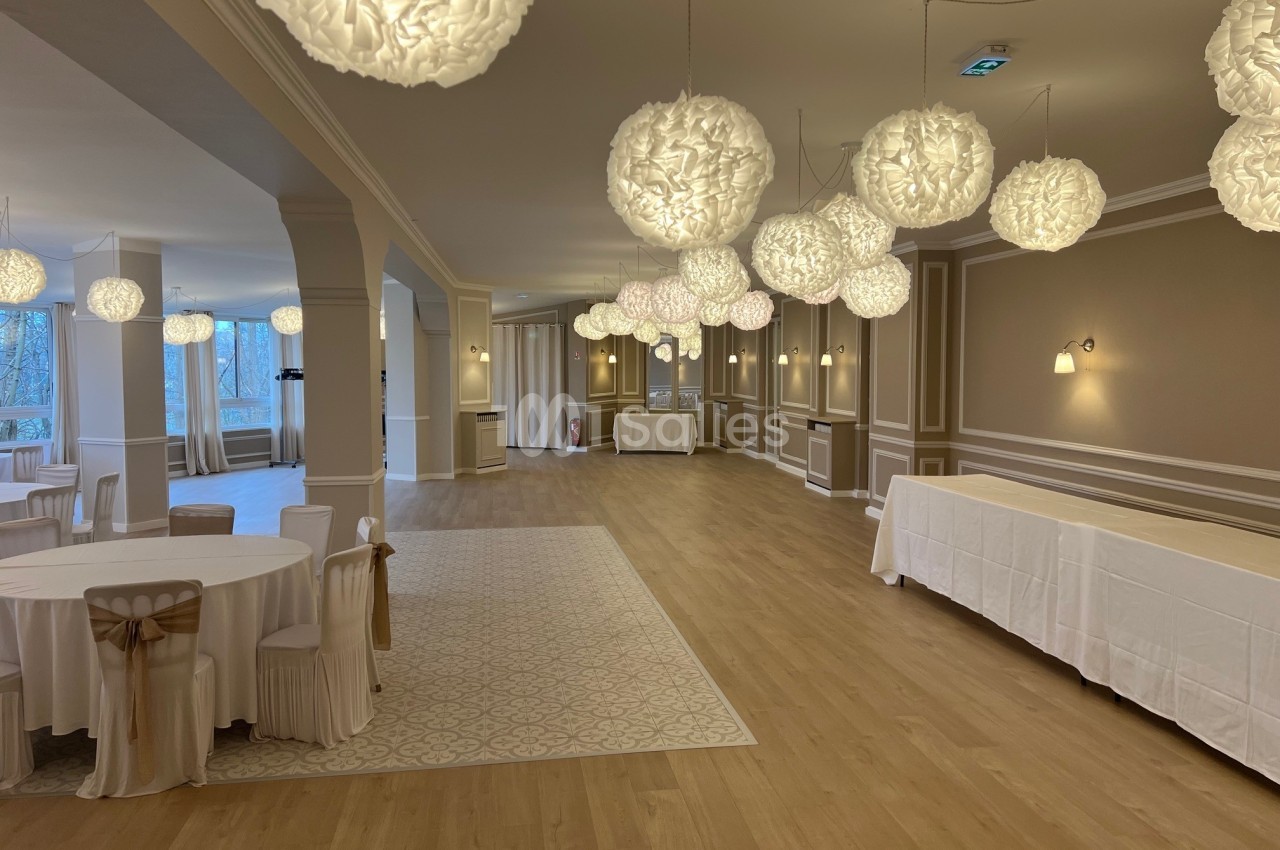 Salle de réception lumineuse avec tables rondes dressées et suspensions décoratives blanches au plafond.