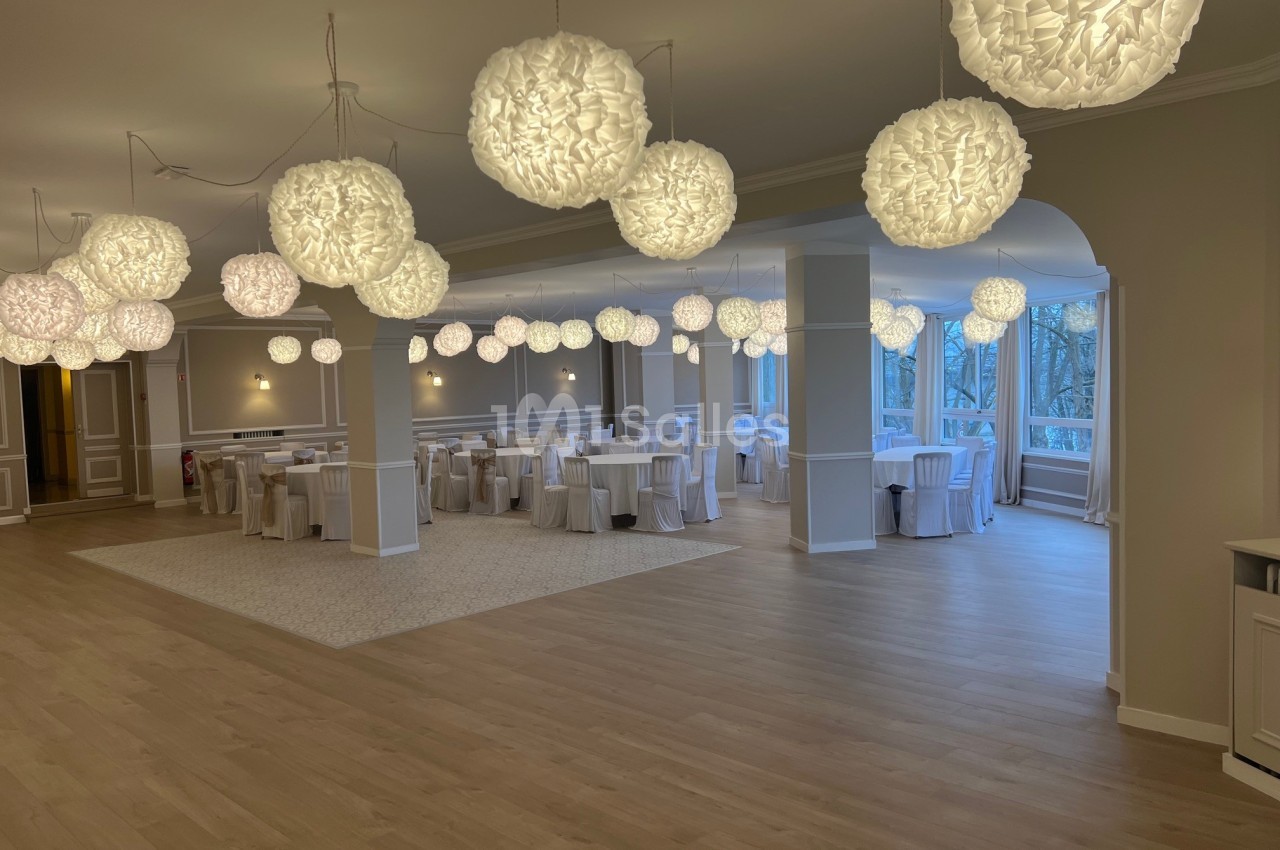 Salle de réception lumineuse avec parquet clair, tables dressées et suspensions décoratives blanches.
