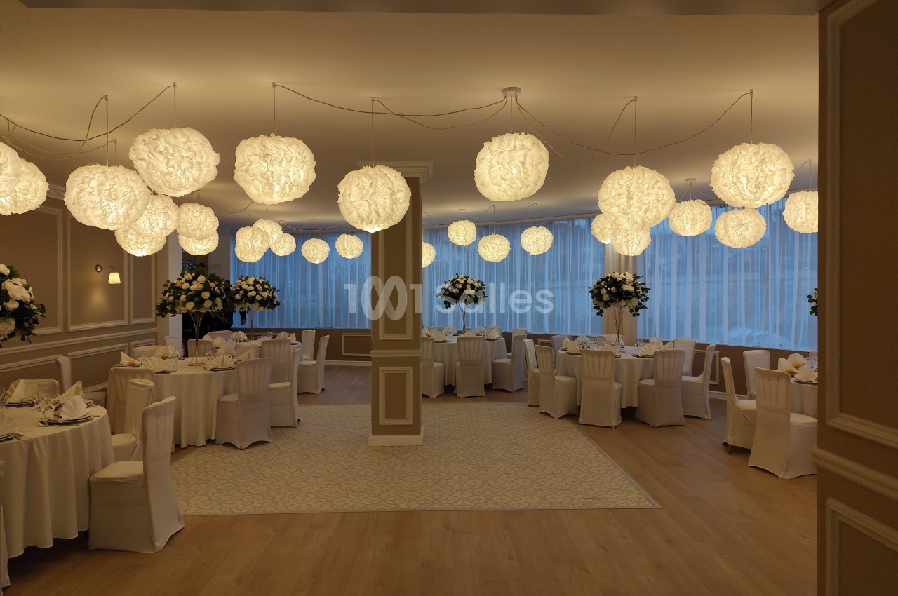 Salle de réception élégante avec tables rondes dressées, chaises blanches et suspensions lumineuses décoratives.