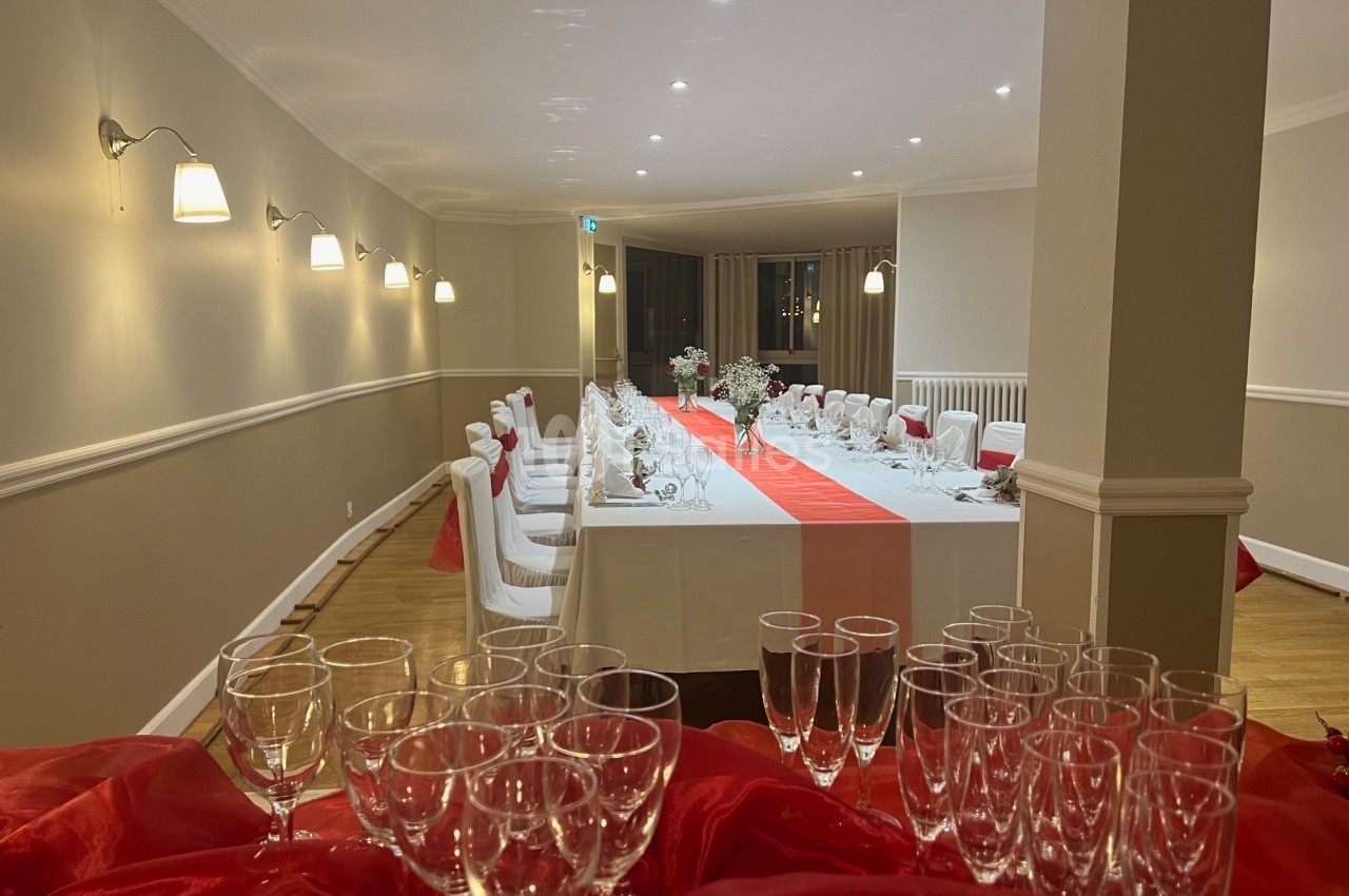 Salle de réception décorée avec une longue table dressée, nappe blanche et chemin de table rouge, verres en premier plan.
