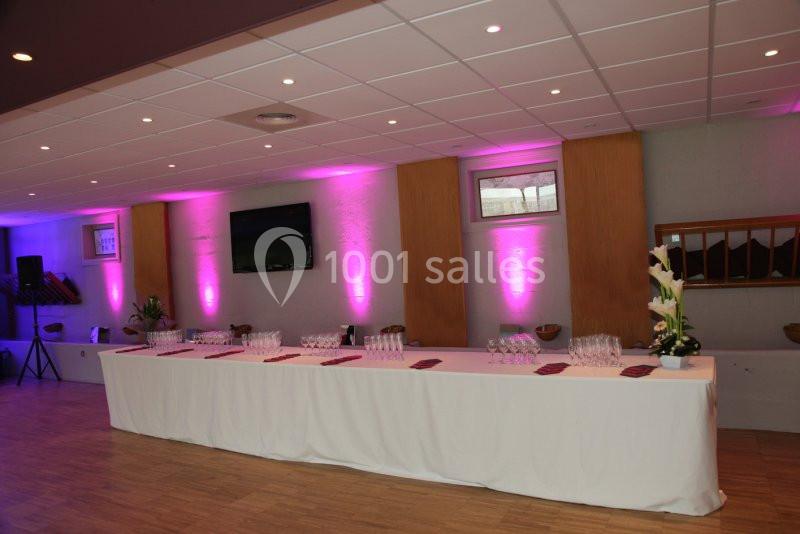 Grande table rectangulaire dressée avec nappes blanches, verres et serviettes, dans une salle éclairée par des lumières…