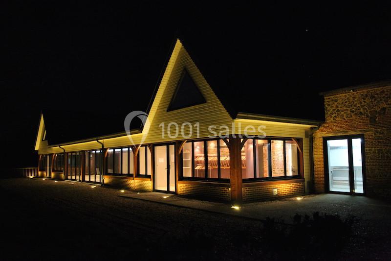 Bâtiment moderne en bois et briques éclairé la nuit, avec de grandes fenêtres laissant entrevoir l'intérieur.