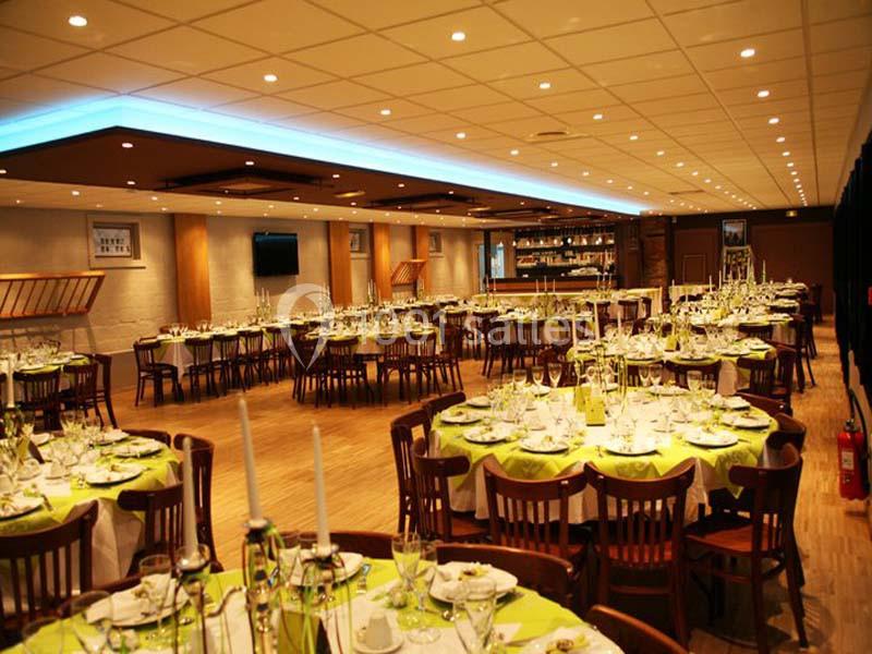 Salle de réception lumineuse avec tables rondes dressées, nappes vertes et chaises en bois, prête pour un événement.