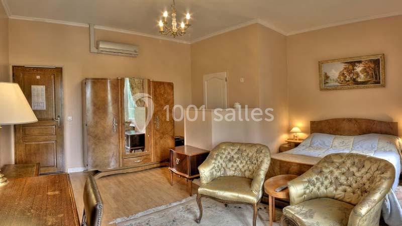 Chambre lumineuse avec lit double, armoire en bois, fauteuils dorés, bureau et éclairage classique.
