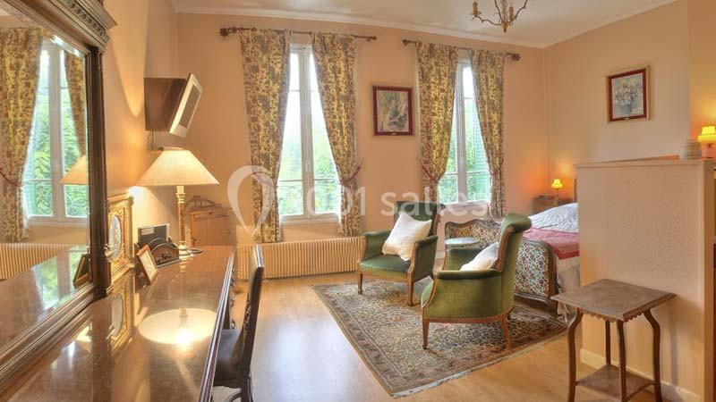 Chambre lumineuse avec lit, fauteuils verts, bureau en bois, rideaux fleuris et vue sur un jardin verdoyant.