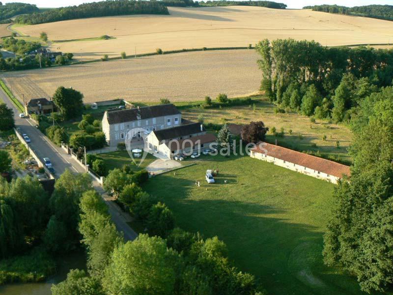 Vue aérienne d'une ferme avec bâtiments, champs cultivés et arbres environnants, située près d'une route de campagne.