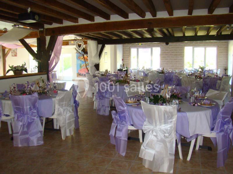 Salle décorée pour un événement avec des tables rondes couvertes de nappes violettes et chaises ornées de nœuds blancs.