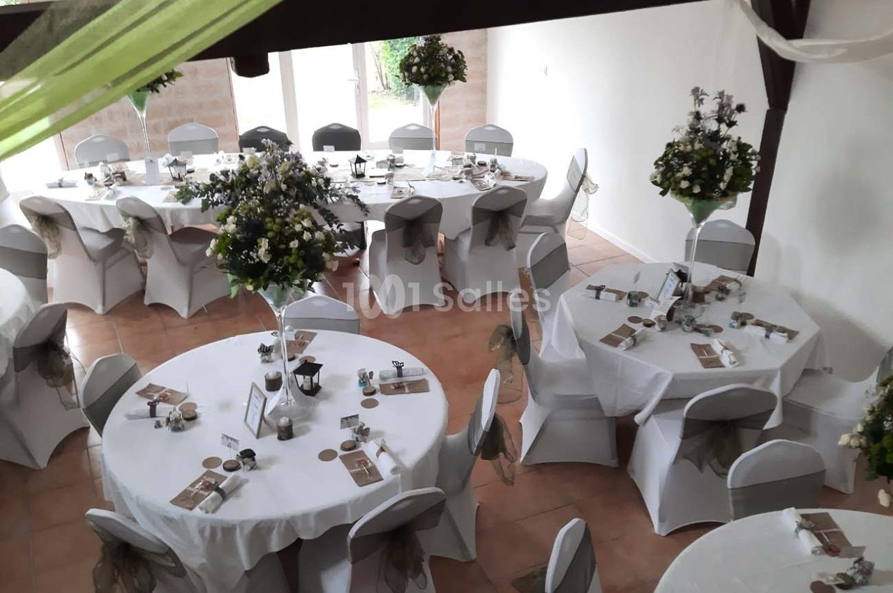 Salle décorée pour un événement avec des tables rondes dressées, nappes blanches et centres de table fleuris.