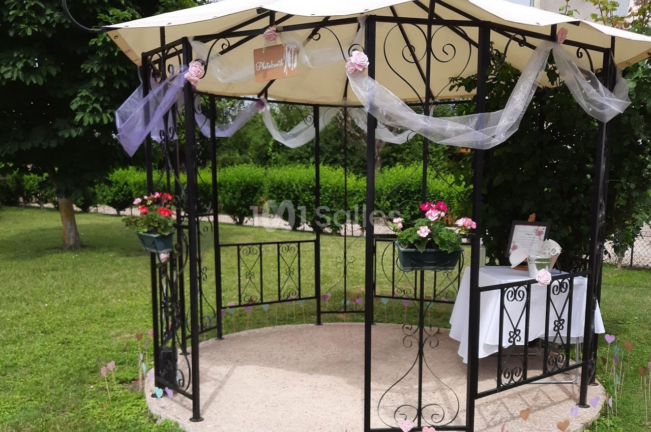 Tonnelle décorée avec des fleurs, voilages et table ornée, installée dans un jardin verdoyant.