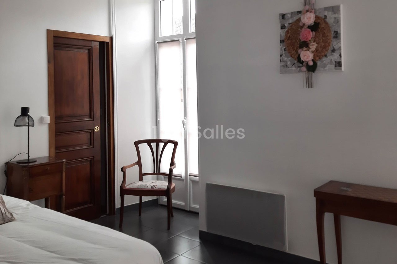Chambre lumineuse avec lit, chaise en bois, bureau, tableau floral au mur et porte en bois foncé.