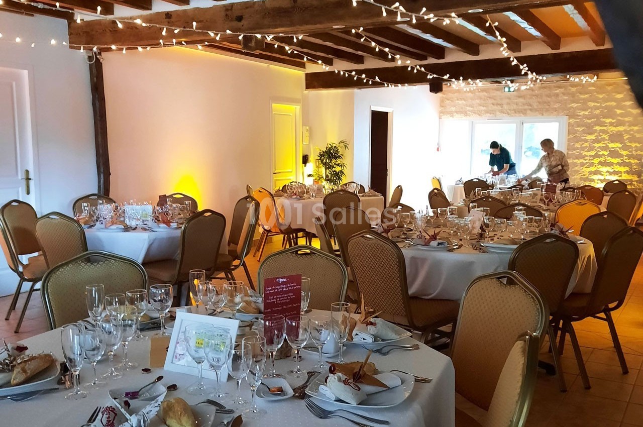 Salle décorée pour un repas festif avec tables rondes dressées, guirlandes lumineuses et deux personnes en préparation.