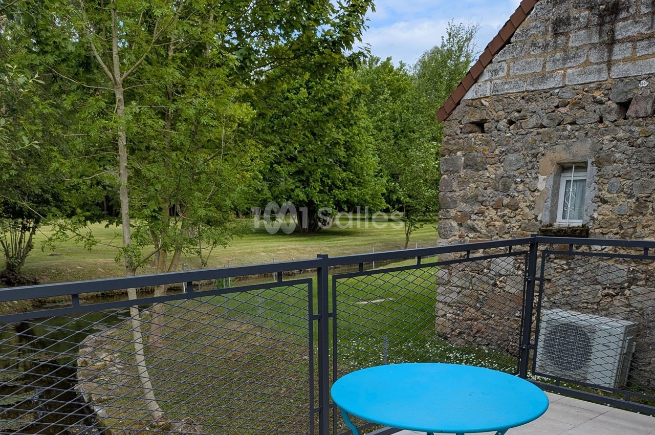 Vue depuis une terrasse avec table bleue, donnant sur un jardin verdoyant et un bâtiment en pierre.