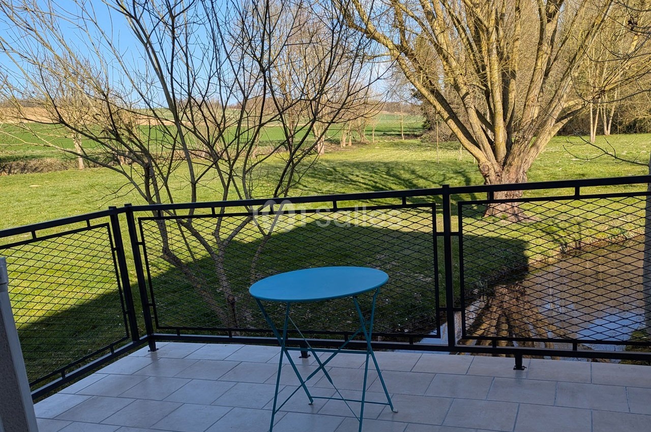 Vue d'une terrasse avec une table bleue, donnant sur un jardin verdoyant avec un arbre et un ruisseau.