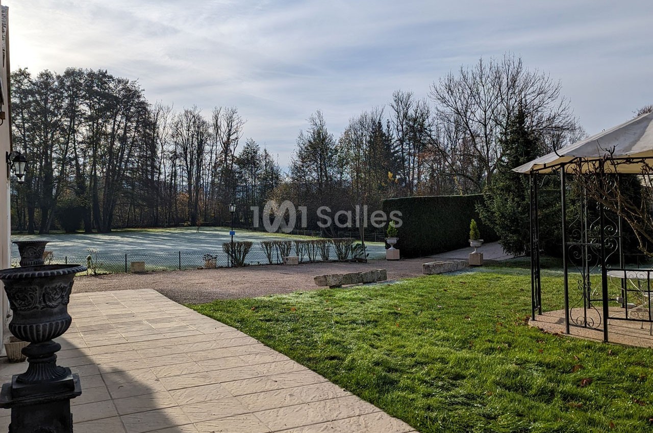 Vue d'un jardin avec pelouse, arbres en arrière-plan, terrasse pavée et pergola métallique sur la droite.