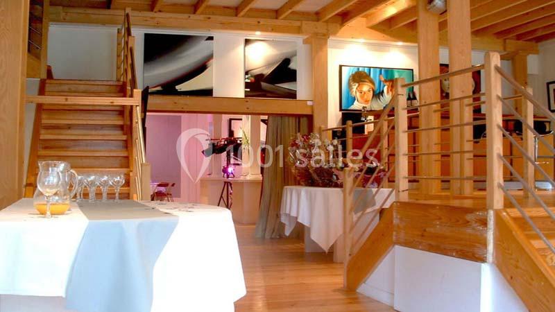 Salle lumineuse avec escalier en bois, tables dressées et décoration florale, ambiance chaleureuse et moderne.