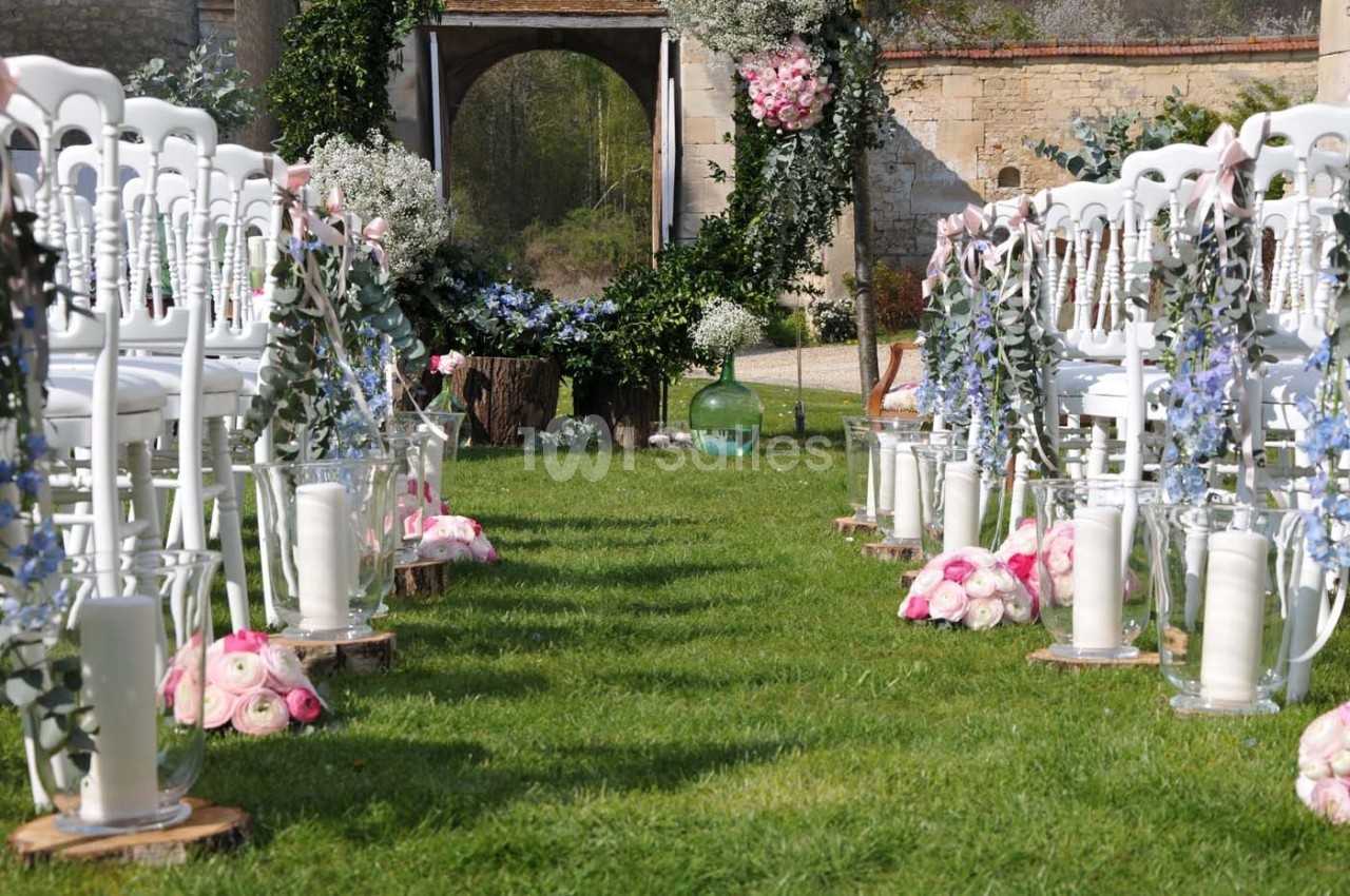 Allée centrale décorée de fleurs et bougies, bordée de chaises blanches, menant à une arche florale en extérieur.