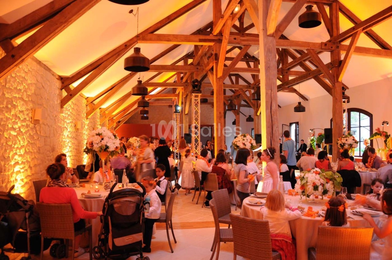 Salle de réception avec des poutres apparentes, décorations florales et invités assis autour de tables rondes.