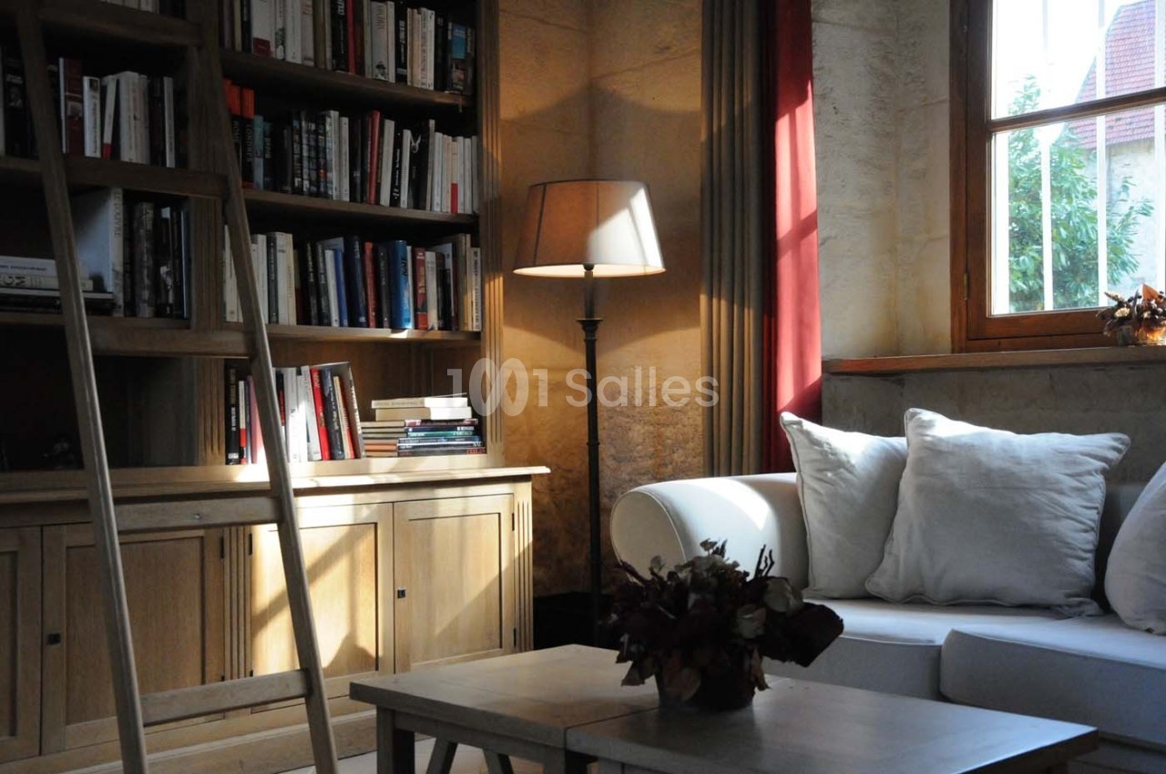 Bibliothèque en bois avec échelle, canapé blanc, lampe allumée et lumière naturelle entrant par une fenêtre.
