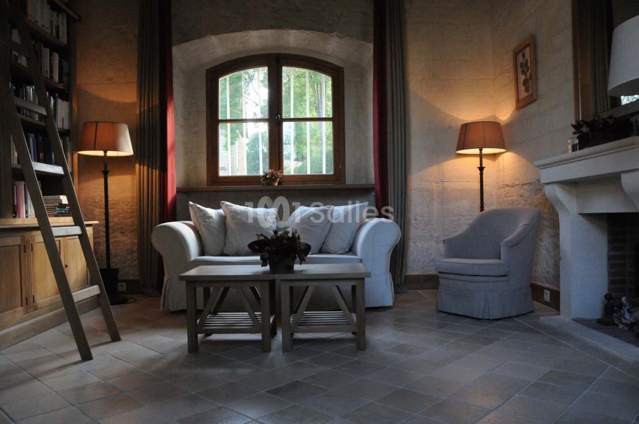 Salon cosy avec canapé blanc, fauteuil gris, table basse en bois, cheminée et lumière douce près d'une fenêtre.