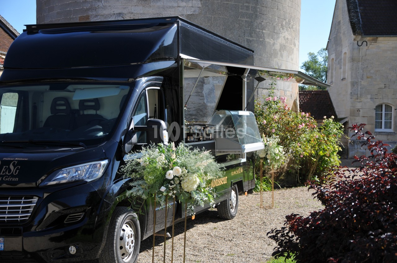 Camion noir aménagé en food truck, décoré de fleurs blanches, stationné devant un bâtiment en pierre.