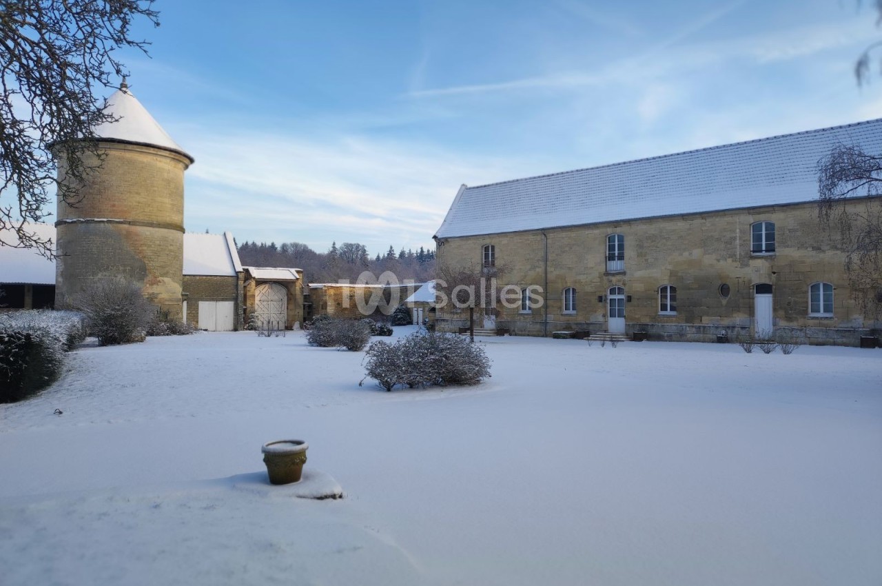 Cour enneigée avec bâtiments en pierre, un pigeonnier cylindrique et des arbres nus sous un ciel dégagé.
