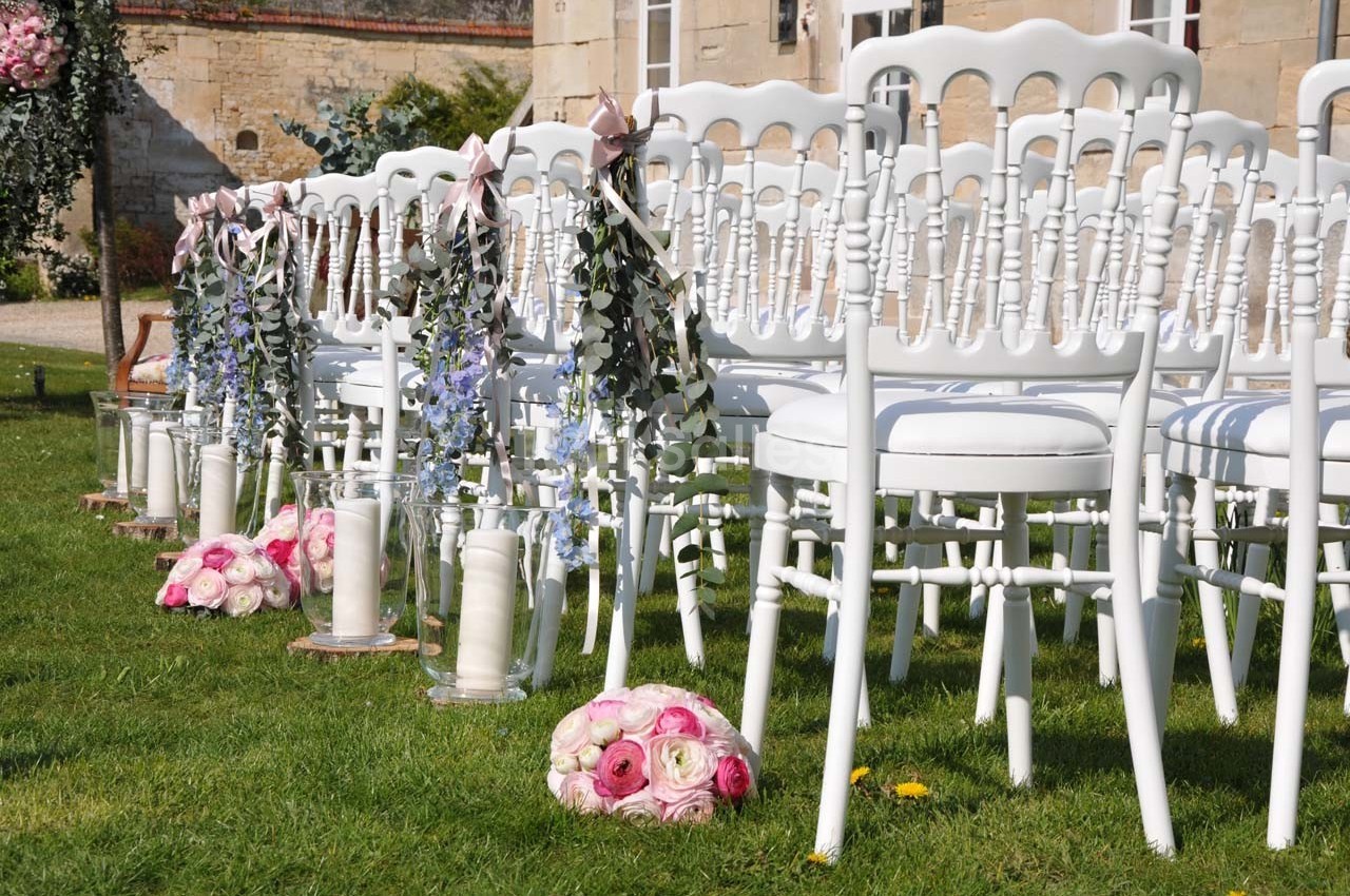Chaises blanches alignées sur une pelouse, décorées de fleurs et de rubans pour une cérémonie en extérieur.