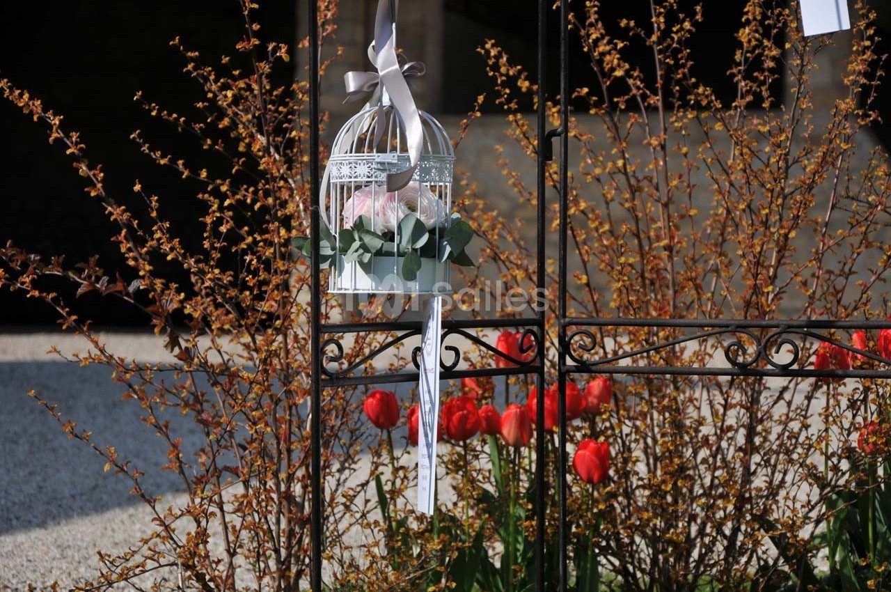 Cage décorative blanche suspendue, contenant des fleurs, devant des tulipes rouges et des branches fleuries.