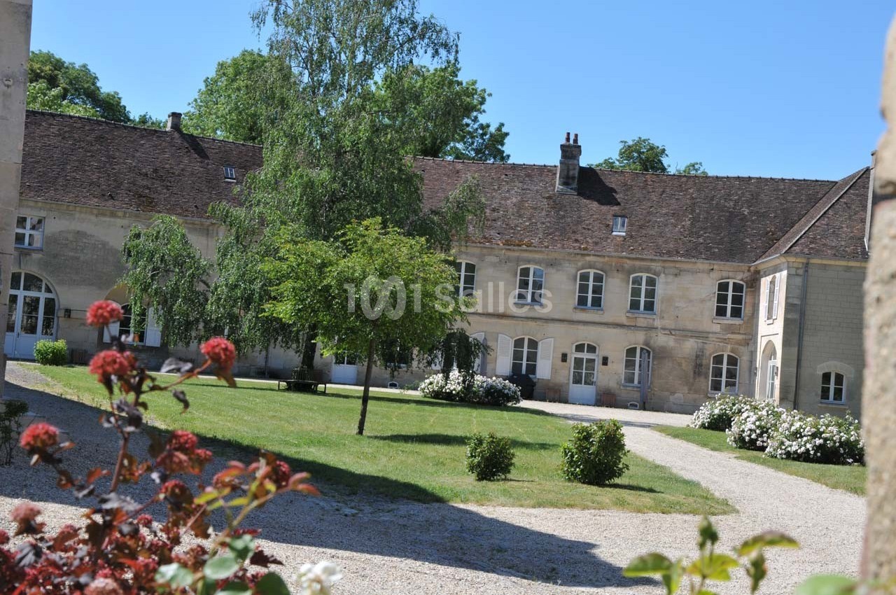 Bâtiment en pierre avec toit en tuiles, entouré d'un jardin verdoyant et de fleurs, sous un ciel dégagé.