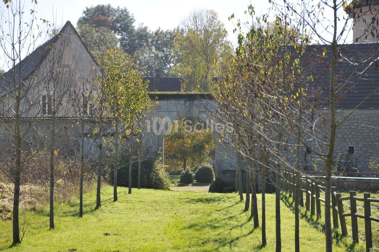 Allée bordée d'arbres menant à une arche en pierre, entourée de bâtiments anciens et de végétation.