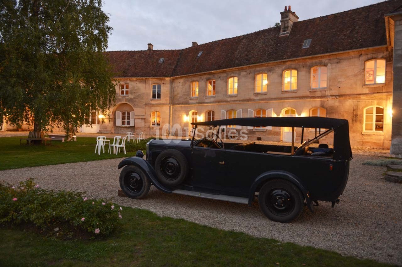 Voiture ancienne noire stationnée dans la cour d'un bâtiment en pierre éclairé à la tombée de la nuit.