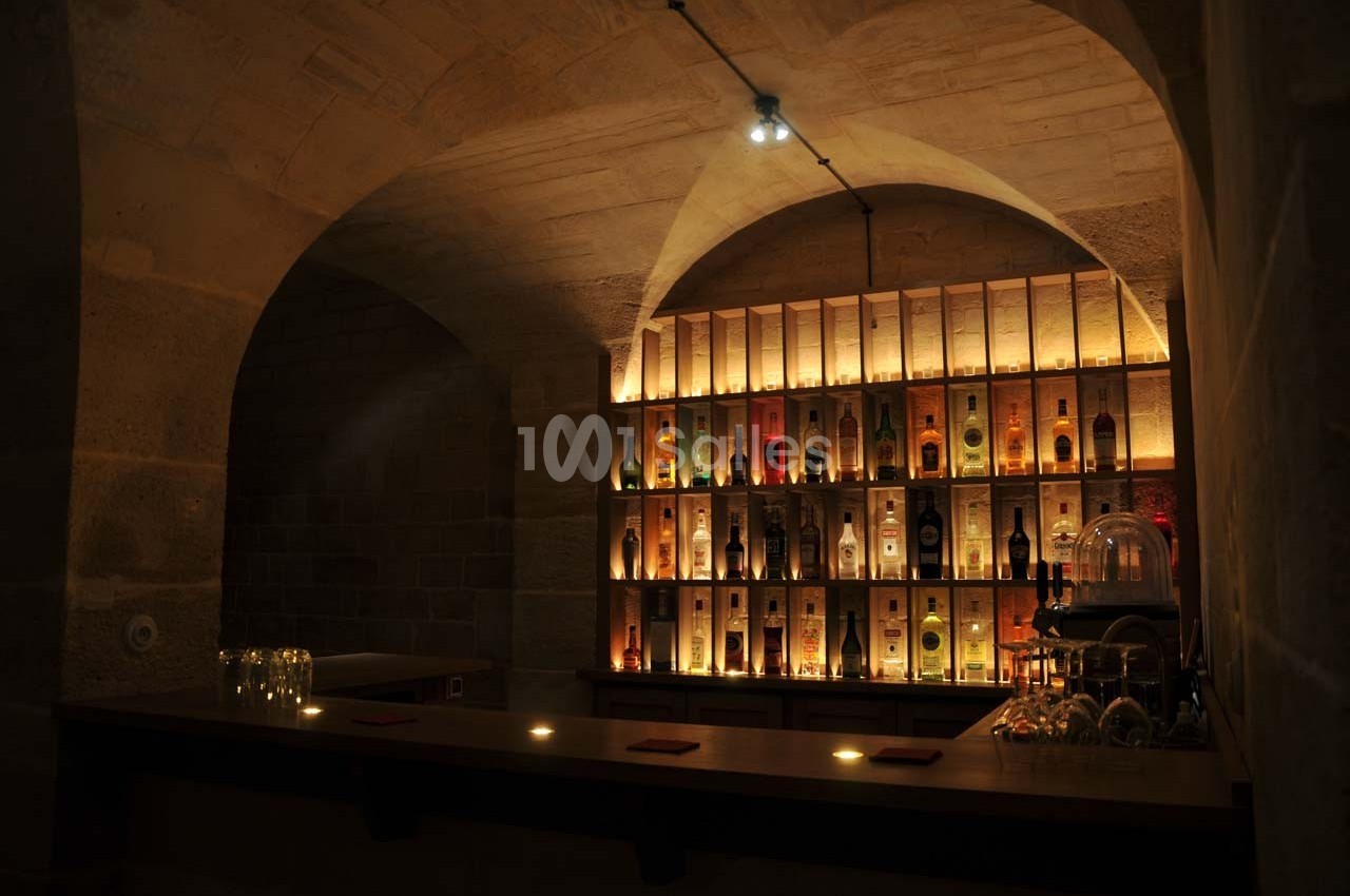 Bar en pierre voûtée avec étagères éclairées exposant des bouteilles d'alcool, ambiance tamisée et chaleureuse.