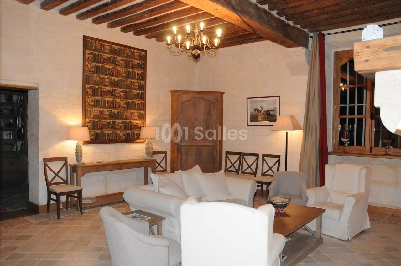 Salon chaleureux avec murs en pierre, poutres apparentes, meubles en bois, fauteuils blancs et éclairage doux.