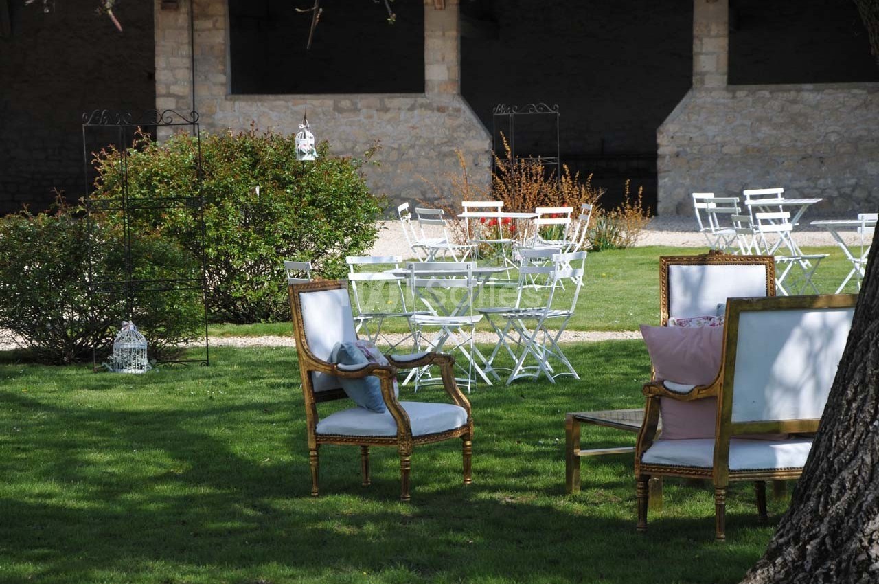 Chaises et fauteuils disposés sur une pelouse, entourés de buissons et de tables blanches dans un jardin extérieur.